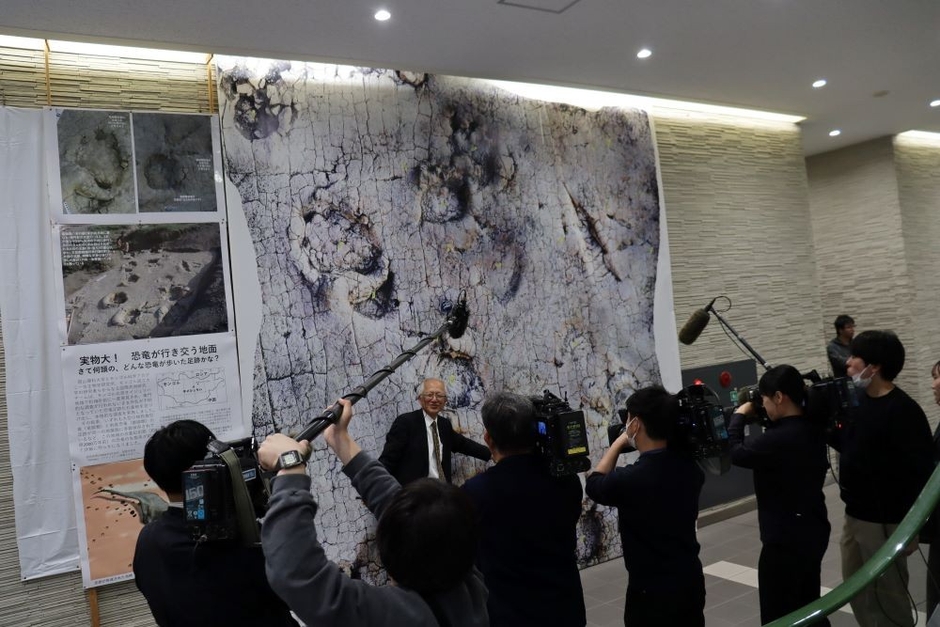 原寸大の足跡化石をプリントしたタペストリーの前で報道陣に解説する石垣名誉館長