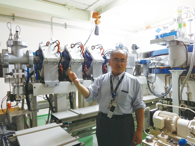 京都大学複合原子力科学研究所内にある世界初のBNCT治療（治験）を実施した加速器の前で解説する鈴木実教授。一つの部屋に入るサイズで、今後病院での運用を可能とする画期的な進歩。