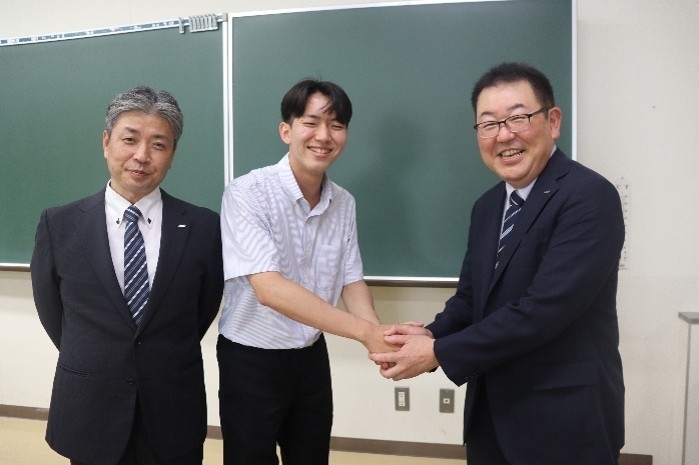 がっちり握手して藤原さん（中央）を激励する烏田社長（右）。左は小林マネージャー