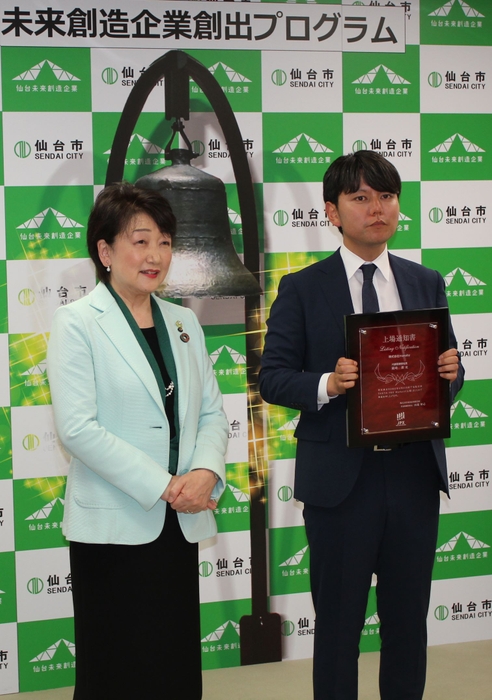 鐘をプリントしたパネルの前で、上場を祝う岡崎社長(右)と郡市長