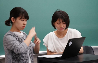 ゼミで手話を使い個人発表する障がいを持つ学生(左)とパソコンでプレゼン原稿の音声化をサポートする学生(右)