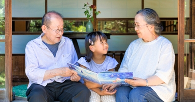 縁側で祖父母と孫がマイステラの絵本を読む様子