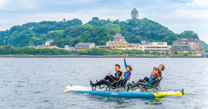 江ノ島の景色をバックに最大4人乗りの足漕ぎペダル式ボードを体験する様子